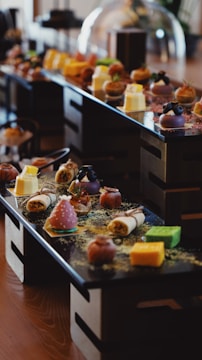 A table adorned with an assortment of intricately crafted desserts and small food items, displayed on multiple tiered black trays. Items include colorful cakes, pastries, and rolled snacks, decorated with fruits and garnishes. The setting appears elegant and thoughtfully arranged, with blurred background elements suggesting a sophisticated venue.