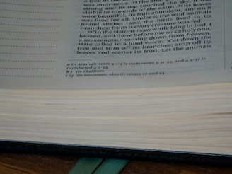 A close-up view of an open book showing printed text on a page, with visible margins and a partially visible other page. The text contains alternating paragraphs with numbers indicating verses and footnotes at the bottom. The book appears to be resting on a wooden surface, with the edge of the pages slightly blurred.