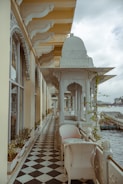 A terrace with an intricate architectural design, featuring ornate arches and decorative columns. The floor is tiled in a black and white checkered pattern, and potted plants line the walls. White wicker chairs and a small table are placed along the corridor, which overlooks a body of water with boats in the distance under a cloudy sky.