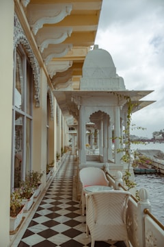 A terrace with an intricate architectural design, featuring ornate arches and decorative columns. The floor is tiled in a black and white checkered pattern, and potted plants line the walls. White wicker chairs and a small table are placed along the corridor, which overlooks a body of water with boats in the distance under a cloudy sky.