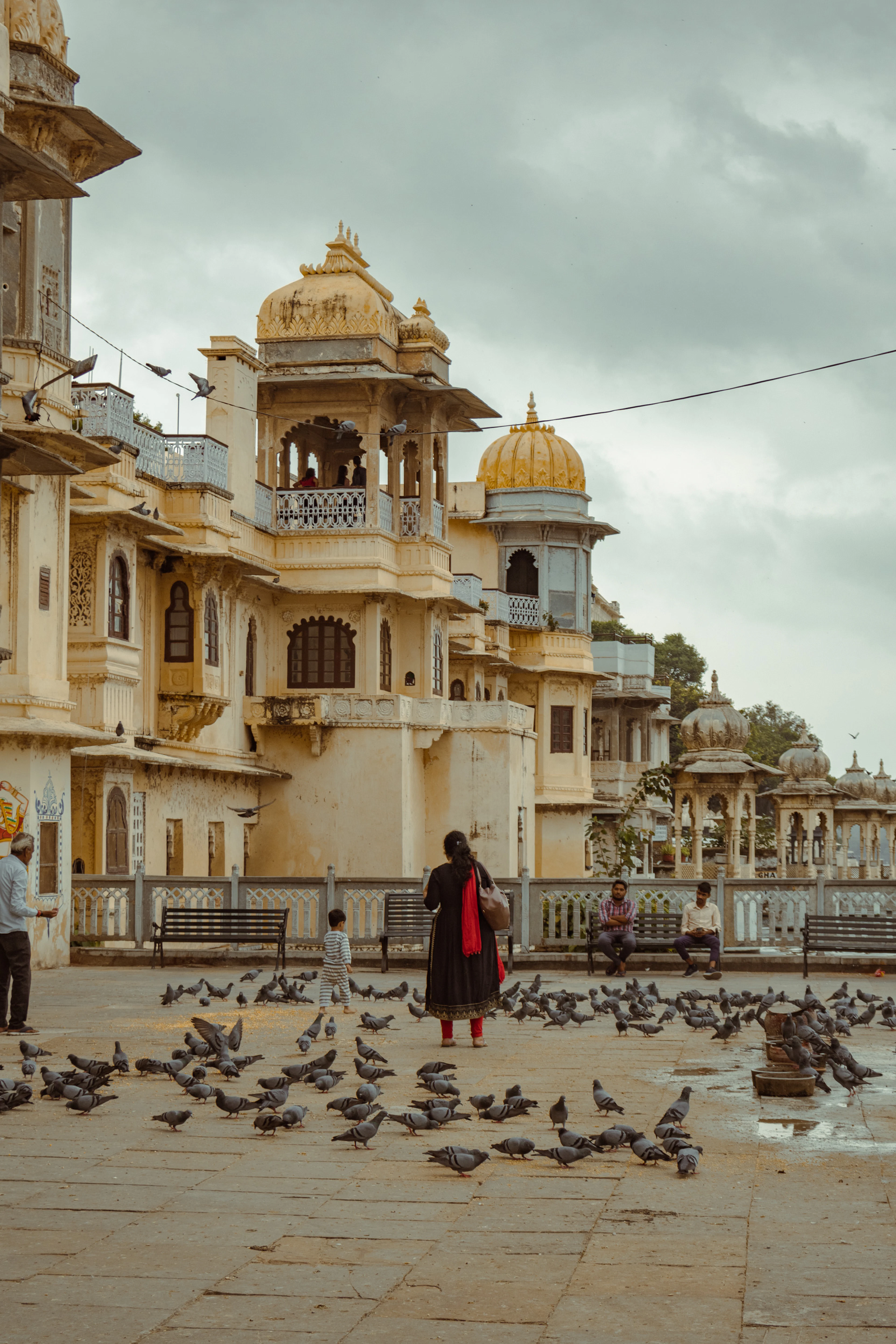 Late afternoon on the stone plaza at Gangaur Ghat in Udaipur: a woman in a red dupatta stands among a scattering of pigeons while a small boy runs past, benches of people watching, the yellow-domed chhatri and carved haveli balconies rising behind under a grey monsoon sky