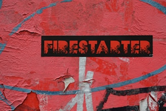 A weathered and peeling red surface forms the background, featuring a bold black sticker with the word 'FIRESTARTER' in red, distressed lettering. The texture and layers of the paint or paper beneath are visibly worn.