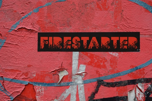 A weathered and peeling red surface forms the background, featuring a bold black sticker with the word 'FIRESTARTER' in red, distressed lettering. The texture and layers of the paint or paper beneath are visibly worn.