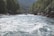A fast-flowing river with white water rapids is surrounded by lush green forests and rocky banks. In the distance, a group of people can be seen rafting down the river. A bridge spans the river further upstream, against a backdrop of wooded hills.