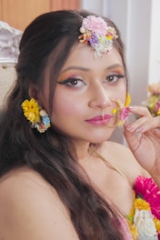 A woman adorned with colorful floral accessories including flowers in her hair and around her ear. Her makeup is vibrant, featuring bold eye shadow and lipstick. She is holding a pink flower close to her face, creating a striking and artistic look.