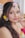 A woman adorned with colorful floral accessories including flowers in her hair and around her ear. Her makeup is vibrant, featuring bold eye shadow and lipstick. She is holding a pink flower close to her face, creating a striking and artistic look.