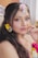A woman adorned with colorful floral accessories including flowers in her hair and around her ear. Her makeup is vibrant, featuring bold eye shadow and lipstick. She is holding a pink flower close to her face, creating a striking and artistic look.