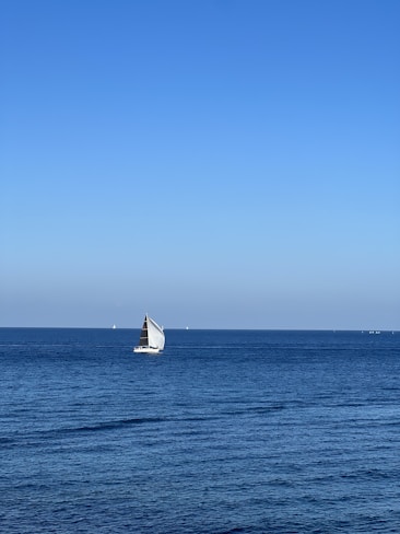 A sailboat with white sails glides smoothly across a vast expanse of calm blue sea under a clear sky. Several distant boats are visible near the horizon, emphasizing the tranquility and open space of the ocean.