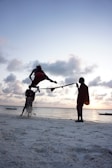 A person is performing a high jump over a rope held by two other individuals on a sandy beach by the sea, with boats in the distance during a sunset. The sky is filled with clouds, and the lighting creates a silhouette effect.