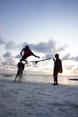 A person is performing a high jump over a rope held by two other individuals on a sandy beach by the sea, with boats in the distance during a sunset. The sky is filled with clouds, and the lighting creates a silhouette effect.