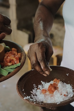 A close-up of hands preparing a dish, with one hand holding a bowl containing shredded coconut, diced tomatoes, and a pinch of spices. Another hand is nearby, holding another bowl filled with chopped tomatoes and green leaves.