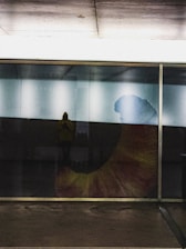 A large glass wall reflects interior lighting and faintly reveals a person standing inside. The prominent feature is a large, colorful graphic or artwork depicting a bent shape, possibly a horn or abstract form, with tones of orange, yellow, and blue.
