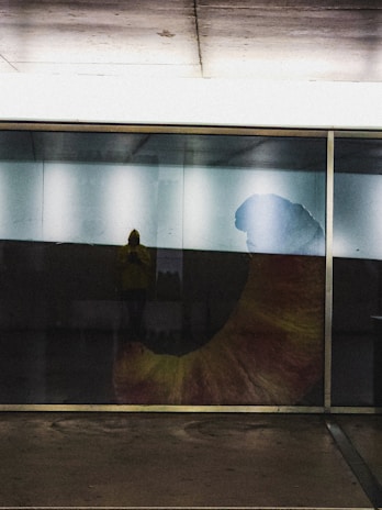 A large glass wall reflects interior lighting and faintly reveals a person standing inside. The prominent feature is a large, colorful graphic or artwork depicting a bent shape, possibly a horn or abstract form, with tones of orange, yellow, and blue.