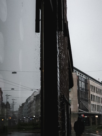 A moody urban scene with a silhouette of a person in the foreground, walking along a rain-soaked street. The image captures towering buildings on either side with reflections from the moisture creating a mirrored effect on the pavement. Overhead, electric wires and street lamps line the street, adding to the overcast ambiance.
