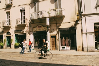 A city street with a group of people gathered near the entrance of a bar labeled 'Bar Milano'. A man rides a bicycle past these people. The buildings have European architectural styles, with balconies and storefronts. Mannequins are displayed in a window of one shop, and green plants adorn the street.