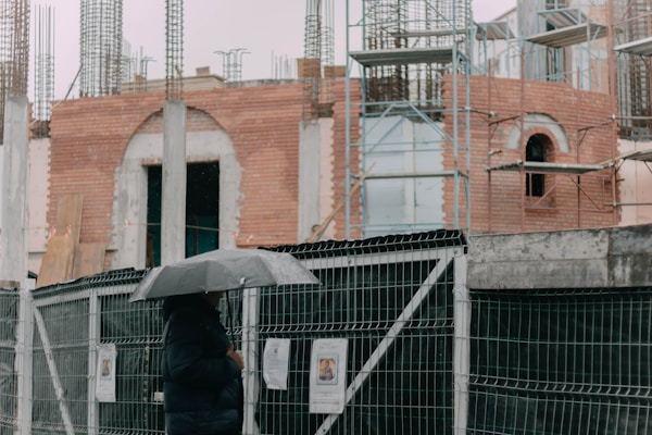 A person holding an umbrella stands in front of a construction site on a rainy day. The site is surrounded by a protective fence, and the building under construction appears to be made of bricks and concrete. Metal reinforcements and scaffolding can be seen, indicating ongoing construction work.