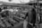 A bustling market scene with several stalls lined up, displaying floral arrangements and various goods. Wooden crates and tables are used to organize and present the merchandise. Multiple people are present, including a person wearing a leather jacket holding a plastic bag, indicating shopping activity. Stalls are covered with branded canopies, providing shelter and a sense of an organized marketplace.