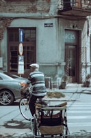An elderly person wearing a cap and striped shirt rides a bicycle with a cart attached at the back, moving along a street with parked cars and a building in the background. The building shows signs of wear, with peeling paint and wires overhead. A street sign indicates a speed limit of 30 and a direction arrow points upwards.