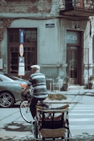 An elderly person wearing a cap and striped shirt rides a bicycle with a cart attached at the back, moving along a street with parked cars and a building in the background. The building shows signs of wear, with peeling paint and wires overhead. A street sign indicates a speed limit of 30 and a direction arrow points upwards.