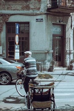 An elderly person wearing a cap and striped shirt rides a bicycle with a cart attached at the back, moving along a street with parked cars and a building in the background. The building shows signs of wear, with peeling paint and wires overhead. A street sign indicates a speed limit of 30 and a direction arrow points upwards.
