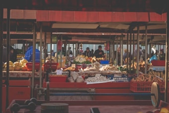 A bustling outdoor market scene filled with various stalls displaying a wide range of fresh produce. The market is shaded by red canopy structures, and numerous people are mingling around the stalls, examining and buying goods. Diverse vegetables like carrots, garlic, potatoes, and other fresh produce are arranged in baskets and trays across the tables.