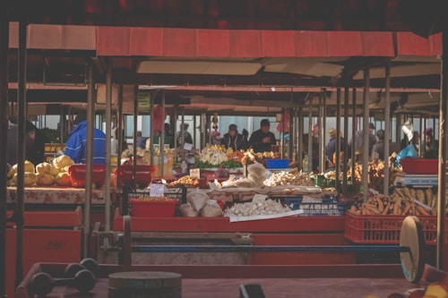 A bustling outdoor market scene filled with various stalls displaying a wide range of fresh produce. The market is shaded by red canopy structures, and numerous people are mingling around the stalls, examining and buying goods. Diverse vegetables like carrots, garlic, potatoes, and other fresh produce are arranged in baskets and trays across the tables.