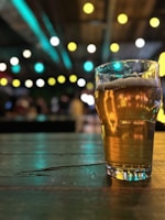 A pint glass filled with beer sits on a wooden table. The background is softly blurred, accented by out-of-focus colorful lights, creating a bokeh effect.