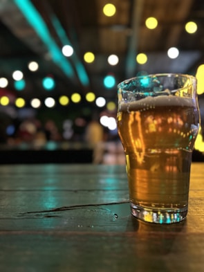 A pint glass filled with beer sits on a wooden table. The background is softly blurred, accented by out-of-focus colorful lights, creating a bokeh effect.