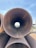Several large metal pipes are stacked on top of each other, with one pipe aligned horizontally pointing directly at the camera. The background is visible through the pipe's circular opening, including a blue sky and some industrial or construction equipment. A person wearing a blue hat can be seen in the distance through the pipe's opening.