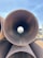 Several large metal pipes are stacked on top of each other, with one pipe aligned horizontally pointing directly at the camera. The background is visible through the pipe's circular opening, including a blue sky and some industrial or construction equipment. A person wearing a blue hat can be seen in the distance through the pipe's opening.
