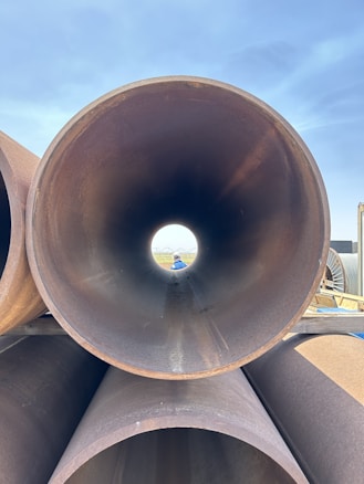 Several large metal pipes are stacked on top of each other, with one pipe aligned horizontally pointing directly at the camera. The background is visible through the pipe's circular opening, including a blue sky and some industrial or construction equipment. A person wearing a blue hat can be seen in the distance through the pipe's opening.