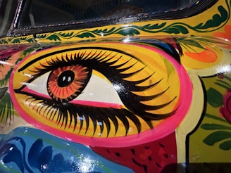 An intricate, colorful painting featuring a large, stylized eye with elaborate eyelashes and vibrant colors surrounding it. The artwork includes detailed patterns and a combination of floral and abstract elements.