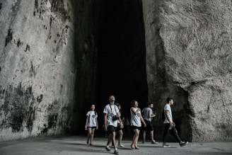A group of people wearing casual clothing walk through a large, cavernous space with high, textured stone walls. The atmosphere is shadowy and cool, with a contrast between the light clothing and the dark interior.