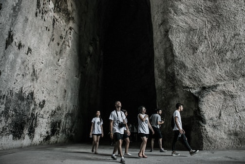 A group of people wearing casual clothing walk through a large, cavernous space with high, textured stone walls. The atmosphere is shadowy and cool, with a contrast between the light clothing and the dark interior.