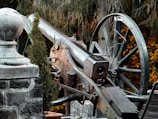 A vintage, historical cannon is situated outdoors, surrounded by greenery. The cannon features a large wheel and rustic metal detailing with visible age and rust. A stone wall and a decorative stone ball are positioned nearby, adding to the historical ambiance. Dense foliage in the background includes autumn-colored leaves, enhancing the scene's picturesque setting.
