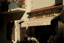 An outdoor view of a building with a beige awning that has 'Made in Sicily' written in red. The facade includes windows with shutters and potted plants. The lighting creates strong shadows, creating a warm, rustic atmosphere.