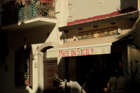 An outdoor view of a building with a beige awning that has 'Made in Sicily' written in red. The facade includes windows with shutters and potted plants. The lighting creates strong shadows, creating a warm, rustic atmosphere.
