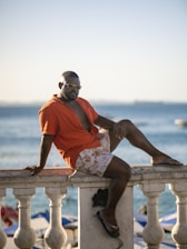 A man with short hair is wearing sunglasses, an unbuttoned orange shirt, patterned shorts, and flip-flops. He is sitting casually on a white railing with ornate designs, near a body of water with boats in the background. The setting is bright and sunny, suggesting a relaxing seaside atmosphere.