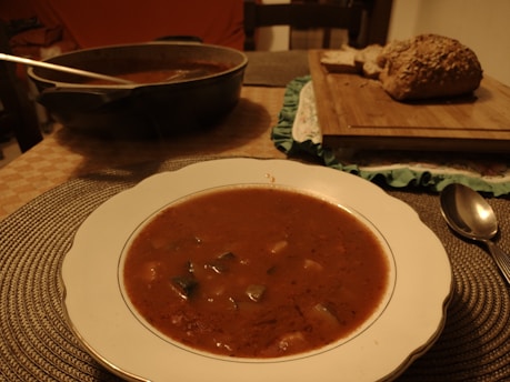 A table setting featuring a bowl of soup with chunks of vegetables, placed on a woven placemat. To the side, there is a spoon and a pot of soup with a ladle. A cutting board holds sliced bread with seeds.