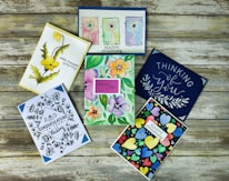 Six colorful greeting cards are arranged on a rustic wooden surface. The cards feature a variety of designs including floral illustrations, abstract heart patterns, and cursive text. Themes include sympathy, congratulations, and love.