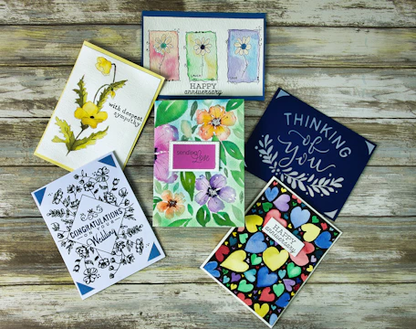 Six colorful greeting cards are arranged on a rustic wooden surface. The cards feature a variety of designs including floral illustrations, abstract heart patterns, and cursive text. Themes include sympathy, congratulations, and love.