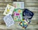 Six colorful greeting cards are arranged on a rustic wooden surface. The cards feature a variety of designs including floral illustrations, abstract heart patterns, and cursive text. Themes include sympathy, congratulations, and love.