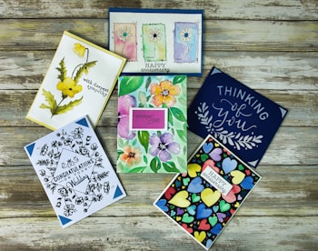 Six colorful greeting cards are arranged on a rustic wooden surface. The cards feature a variety of designs including floral illustrations, abstract heart patterns, and cursive text. Themes include sympathy, congratulations, and love.