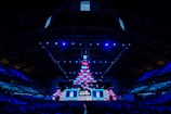 An expansive venue filled with a large audience, featuring a grand stage lit with vibrant blue and pink lights. The ceiling is decorated with a large display, and numerous screens showcase the event's branding. The atmosphere is dynamic and futuristic, reflecting a technologically advanced conference.