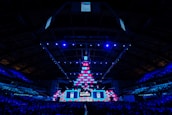An expansive venue filled with a large audience, featuring a grand stage lit with vibrant blue and pink lights. The ceiling is decorated with a large display, and numerous screens showcase the event's branding. The atmosphere is dynamic and futuristic, reflecting a technologically advanced conference.