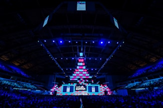 An expansive venue filled with a large audience, featuring a grand stage lit with vibrant blue and pink lights. The ceiling is decorated with a large display, and numerous screens showcase the event's branding. The atmosphere is dynamic and futuristic, reflecting a technologically advanced conference.