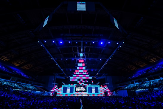 An expansive venue filled with a large audience, featuring a grand stage lit with vibrant blue and pink lights. The ceiling is decorated with a large display, and numerous screens showcase the event's branding. The atmosphere is dynamic and futuristic, reflecting a technologically advanced conference.
