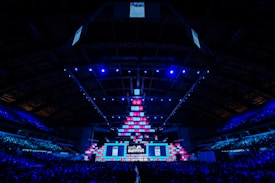 An expansive venue filled with a large audience, featuring a grand stage lit with vibrant blue and pink lights. The ceiling is decorated with a large display, and numerous screens showcase the event's branding. The atmosphere is dynamic and futuristic, reflecting a technologically advanced conference.