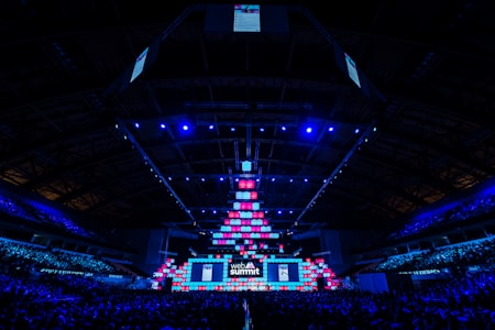 An expansive venue filled with a large audience, featuring a grand stage lit with vibrant blue and pink lights. The ceiling is decorated with a large display, and numerous screens showcase the event's branding. The atmosphere is dynamic and futuristic, reflecting a technologically advanced conference.