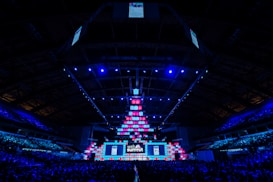 An expansive venue filled with a large audience, featuring a grand stage lit with vibrant blue and pink lights. The ceiling is decorated with a large display, and numerous screens showcase the event's branding. The atmosphere is dynamic and futuristic, reflecting a technologically advanced conference.